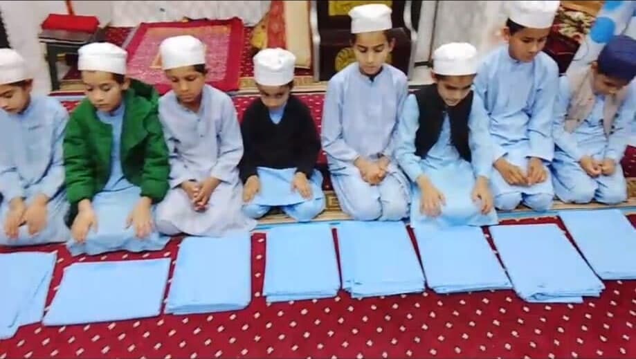 A row of boys in white caps sit on a red carpet, each with a folded set of new clothes