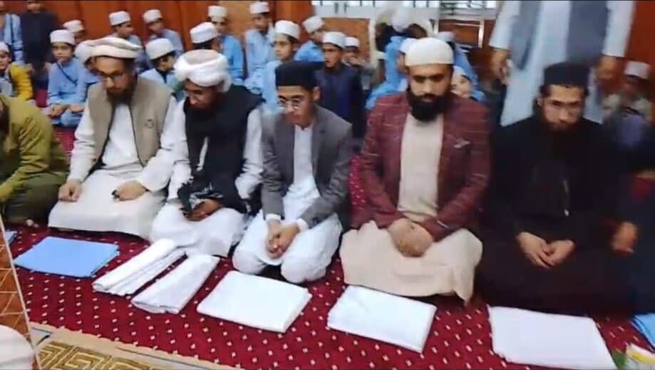 Teachers and rows of students seated on a red carpet with folded uniforms in front of them