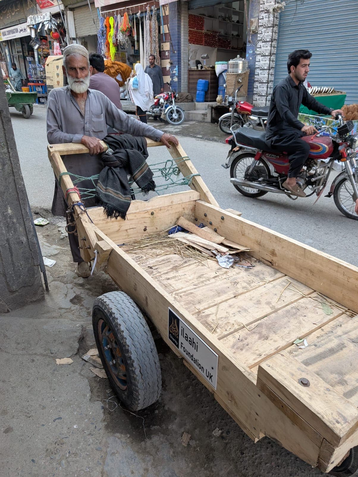 An elderly man sits on a new wooden hand cart labelled Ilaahi Foundation UK on a busy market street in Pakistan