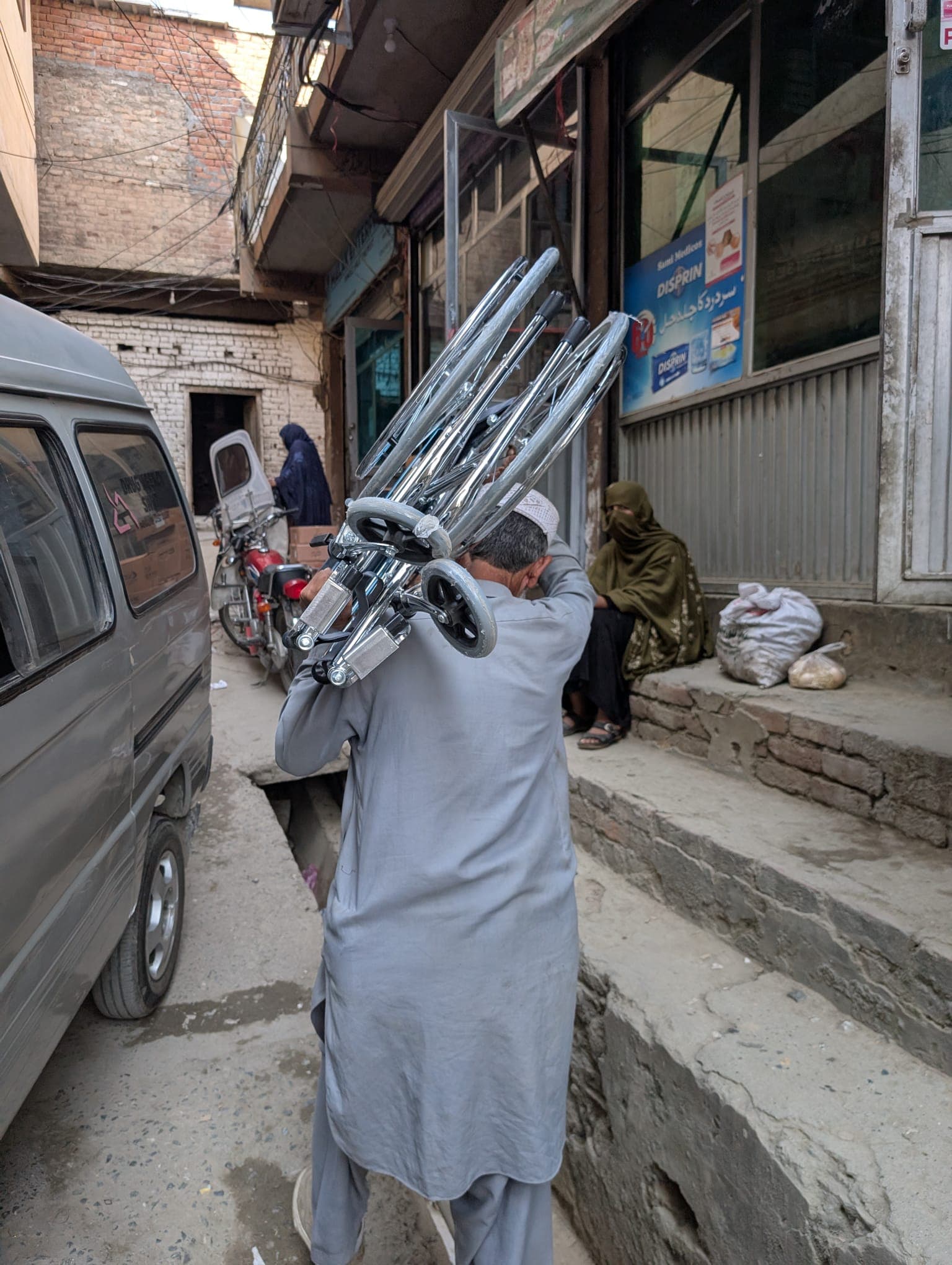 A man carries a stack of folded wheelchairs on his shoulder through a narrow street to deliver them